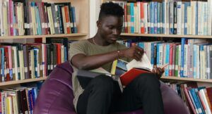 a dark brown-skinned Black man reading in a bean bag in a library